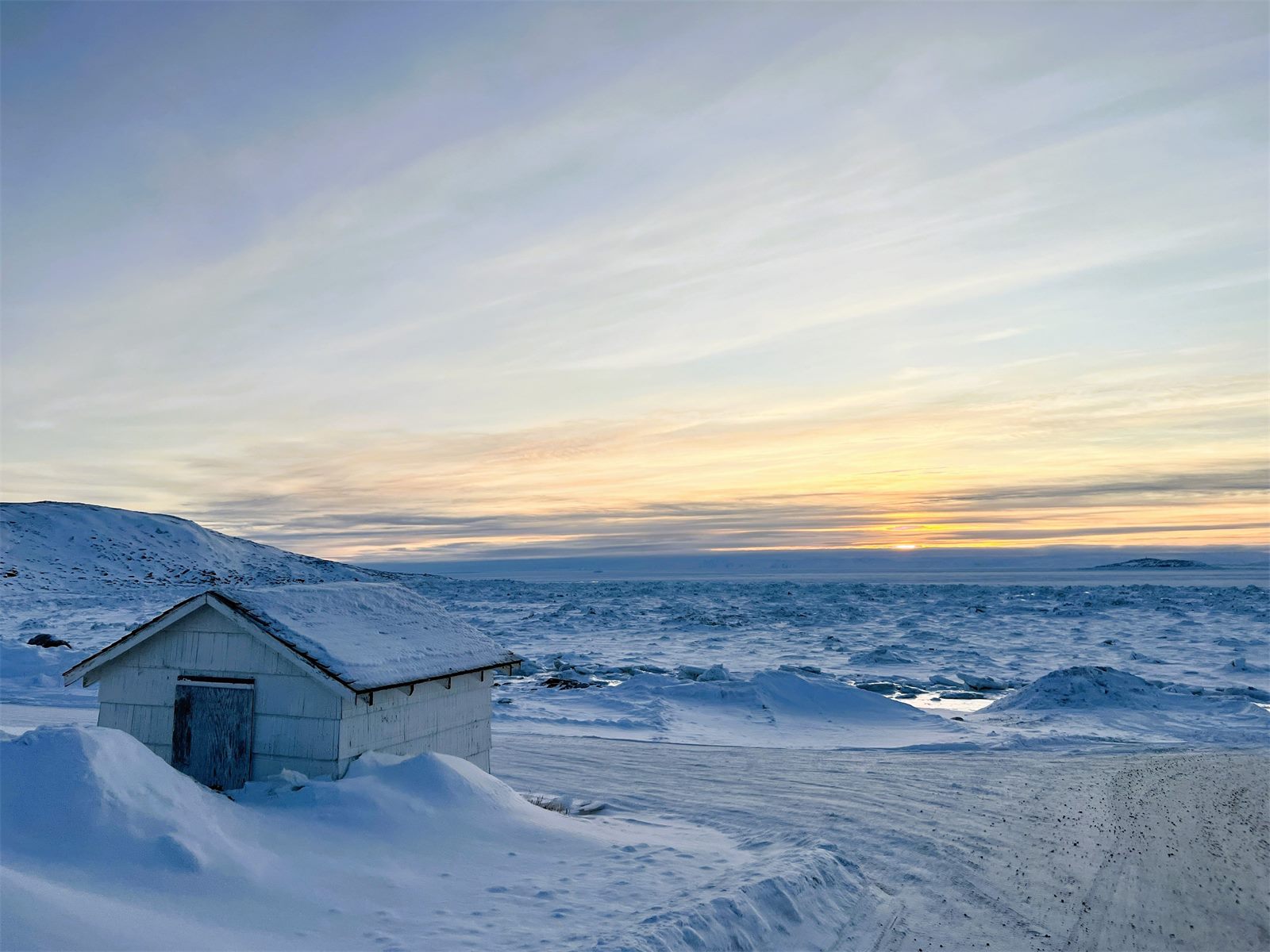 A small snow-covered cabin on a vast frozen Arctic coastline at sunset, with sea ice stretching to the horizon