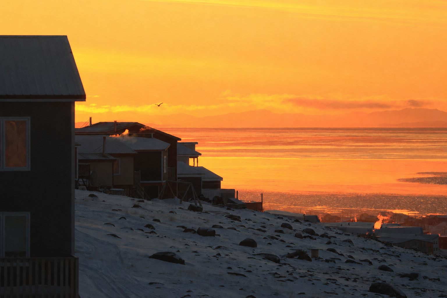 A remote northern Indigenous community of houses on a snowy coastline at sunset, with smoke rising from the chimneys